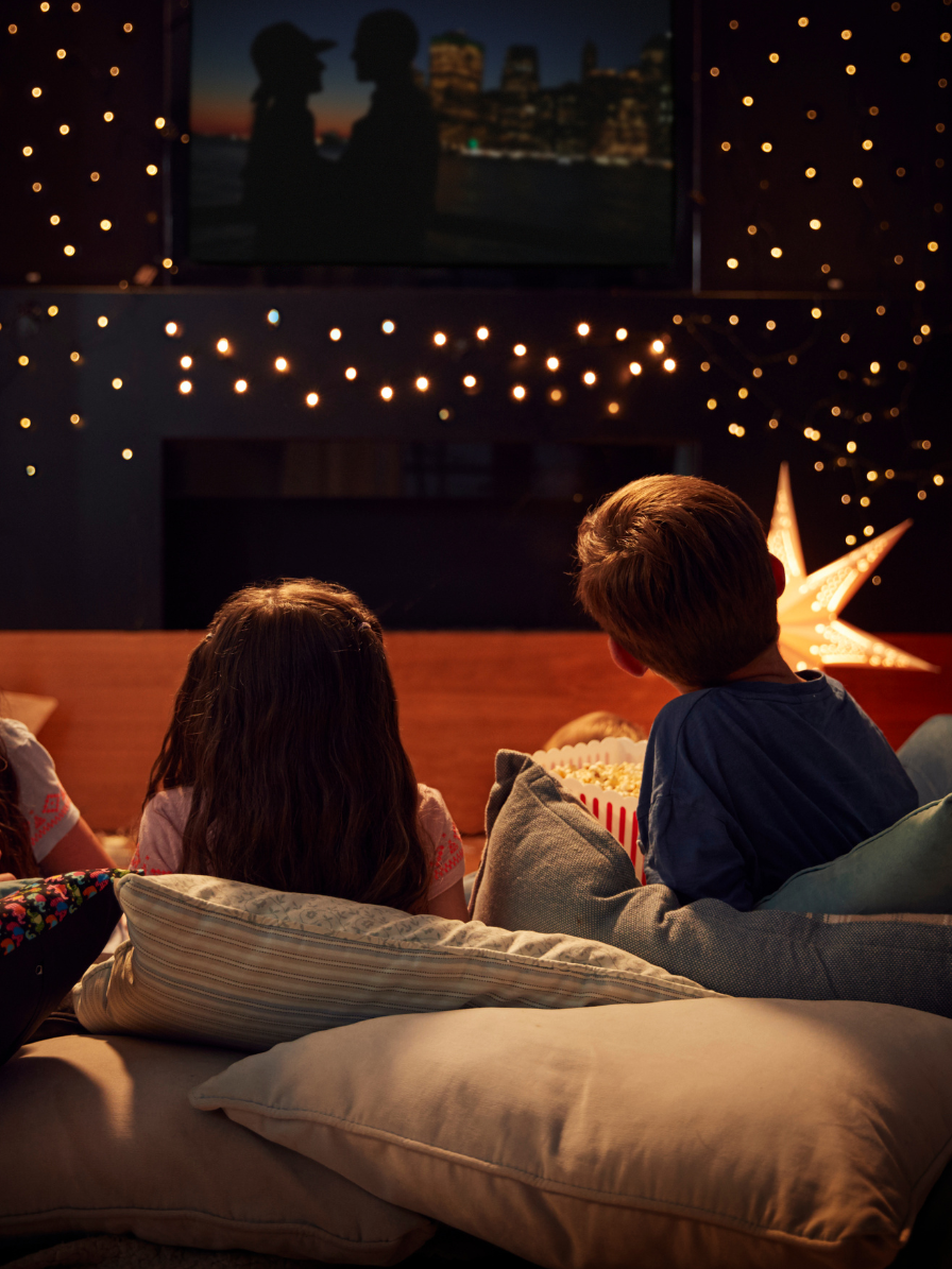 Kids watch a movie on the floor with comfy pillows in a room lit by fairy lights.