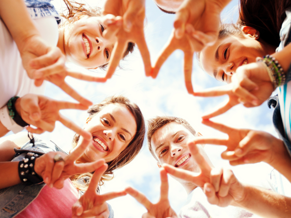 A group of teens standing in a circle, holding up a 2-finger V shape looking down at the camera.