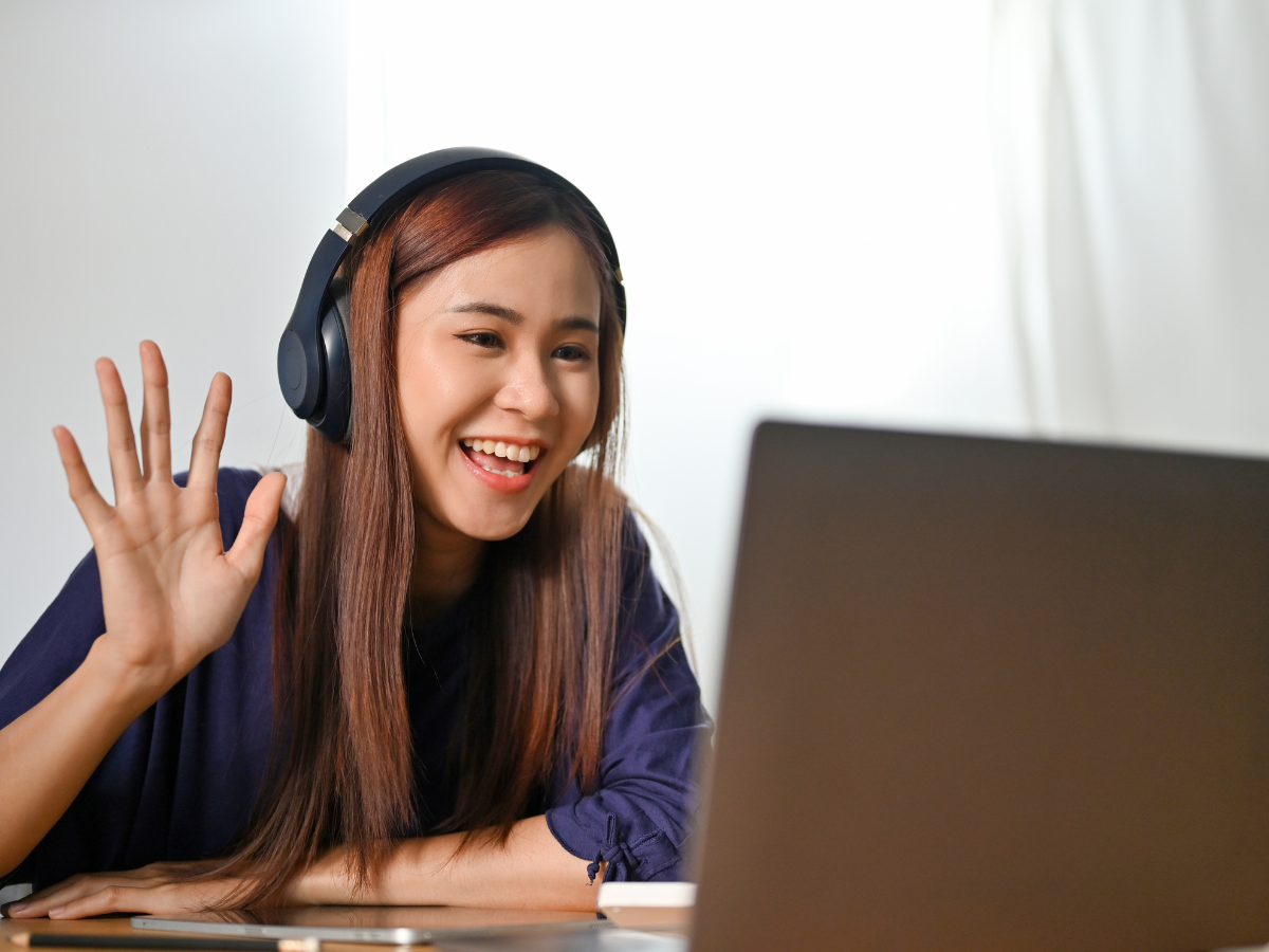 A teen girl wearing headphones and sitting in front of a laptop. She is waving at the screen.