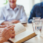 a hand pointing to text in an open book with multiple people visible in the background of the image