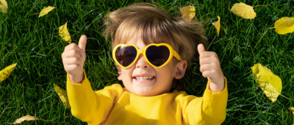 Child laying in the grass wearing yellow heart-shaped sunglasses and smiling while giving two thumbs-up.