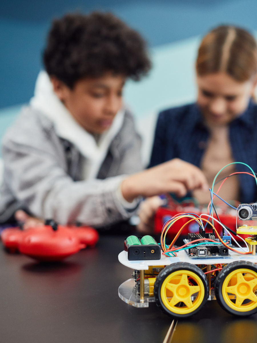 two kids working together to assemble a robot, with small robot on wheels in foreground