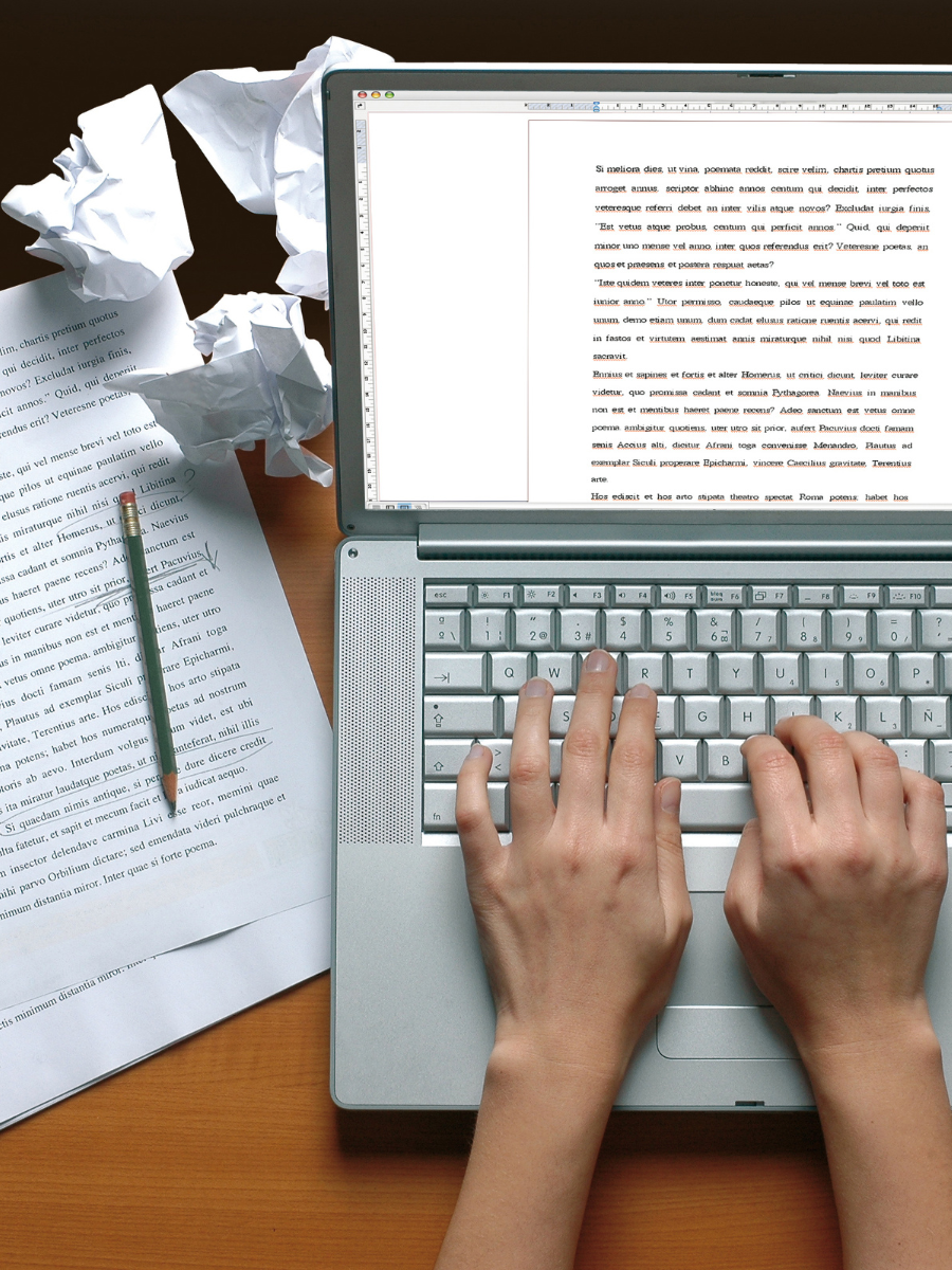 overhead view of hands typing on laptop with paper edits to the right of the computer