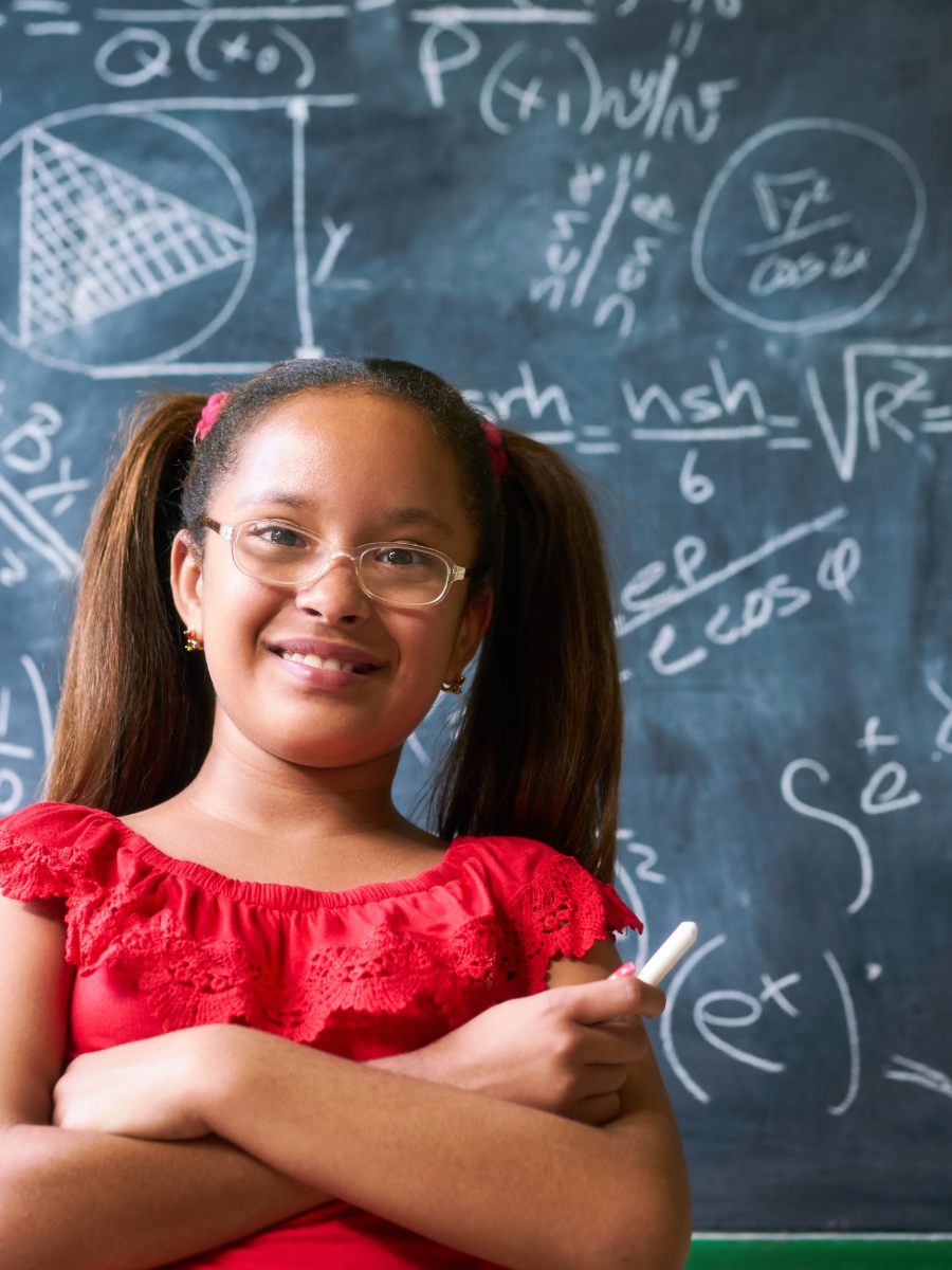 Young girl in pigtails and a red shirt standing in front of a chalkboard with mathematic equations written on it.
