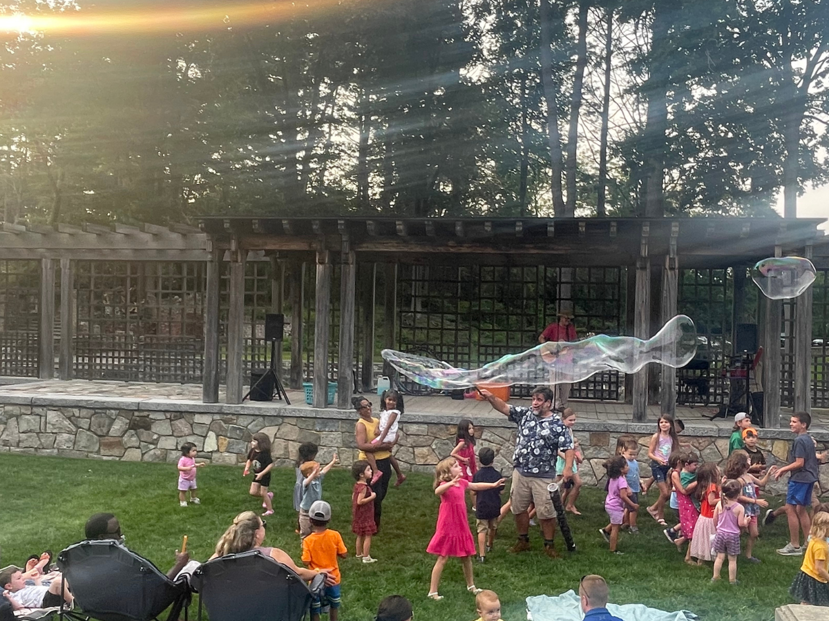 A large, intergenerational group dances and chases giant bubbles in a pretty garden space.