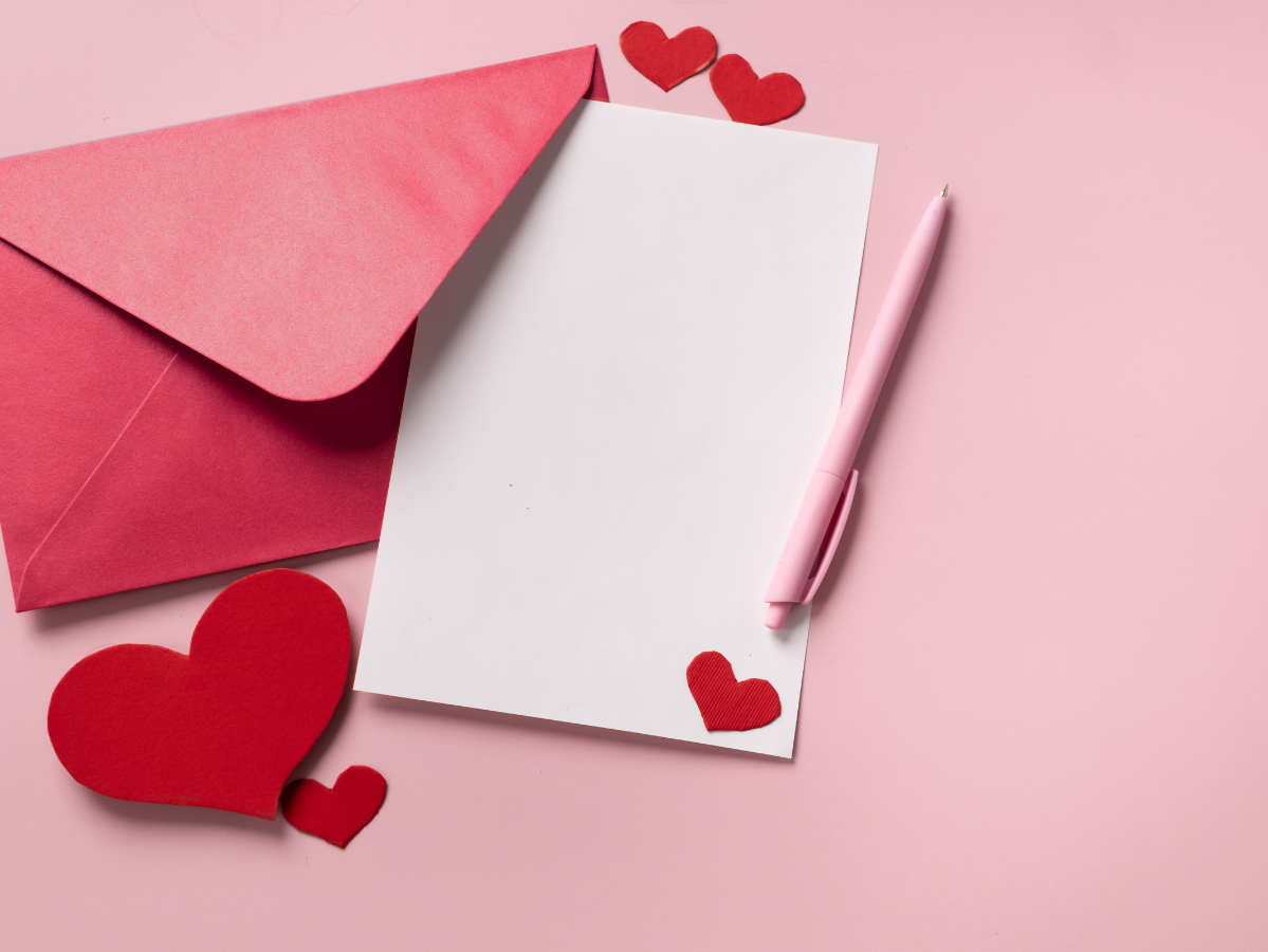 Red envelope and white sheet of paper on pink background with red paper hearts scattered around