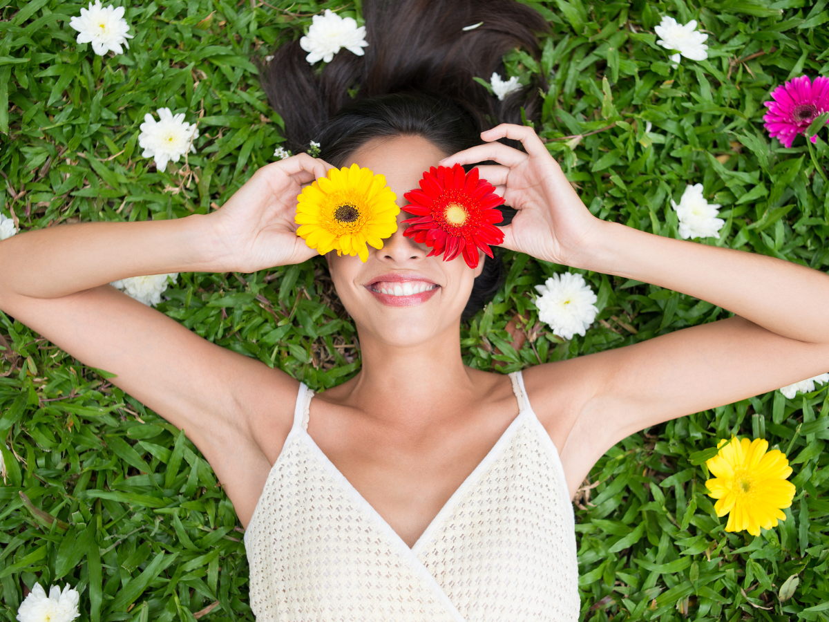 A girl laying in the grass wearing a white sundress holding red and yellow flowers over her eyes.