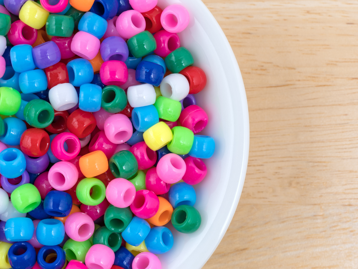 assorted colorful plastic beads in a white bowl on a light brown table.